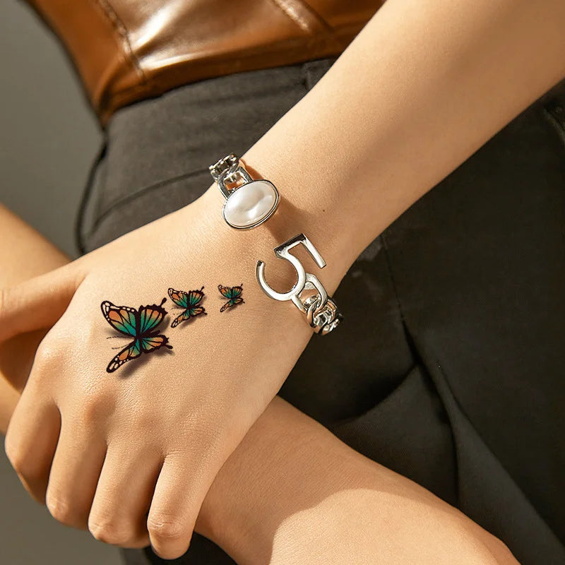 A hand wearing a silver ring and a letter charm with butterfly stickers on the wrist against a dark background.
