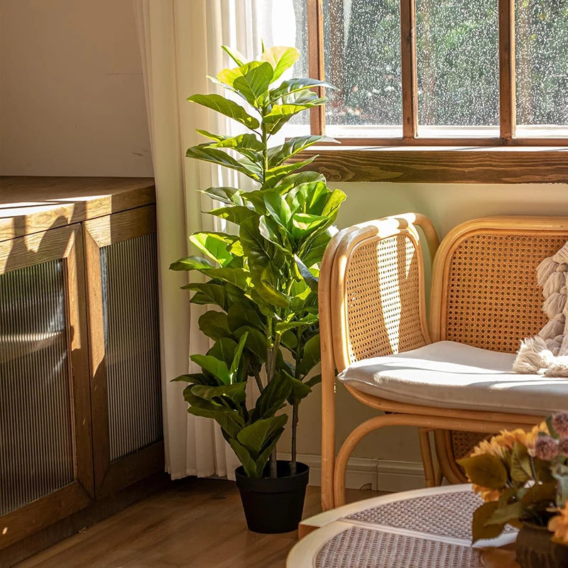 A tall artificial fiddle leaf fig tree in a black pot sits on a wooden shelf next to a light-colored wooden chair.