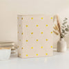 A light beige laminated photo album with gold heart accents sits on a white surface next to a vase of dried flowers.
