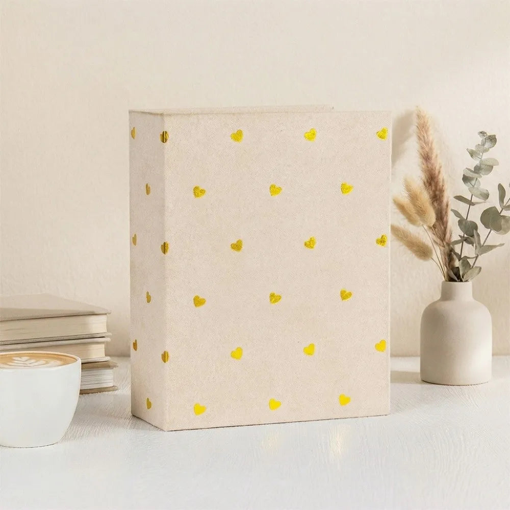 A light beige laminated photo album with gold heart accents sits on a white surface next to a vase of dried flowers.