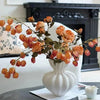 A white ceramic vase filled with dried orange and brown floral elements sits on a dark surface.