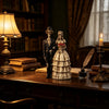 A resin figurine depicting a gothic skeleton bride and groom standing on a wooden desk next to a lamp and candle.