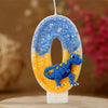 A birthday candle shaped like the number 0, decorated with blue glitter and an orange dinosaur figure.
