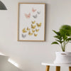 A framed watercolor illustration of various colored butterflies on a white wall above a potted green plant.