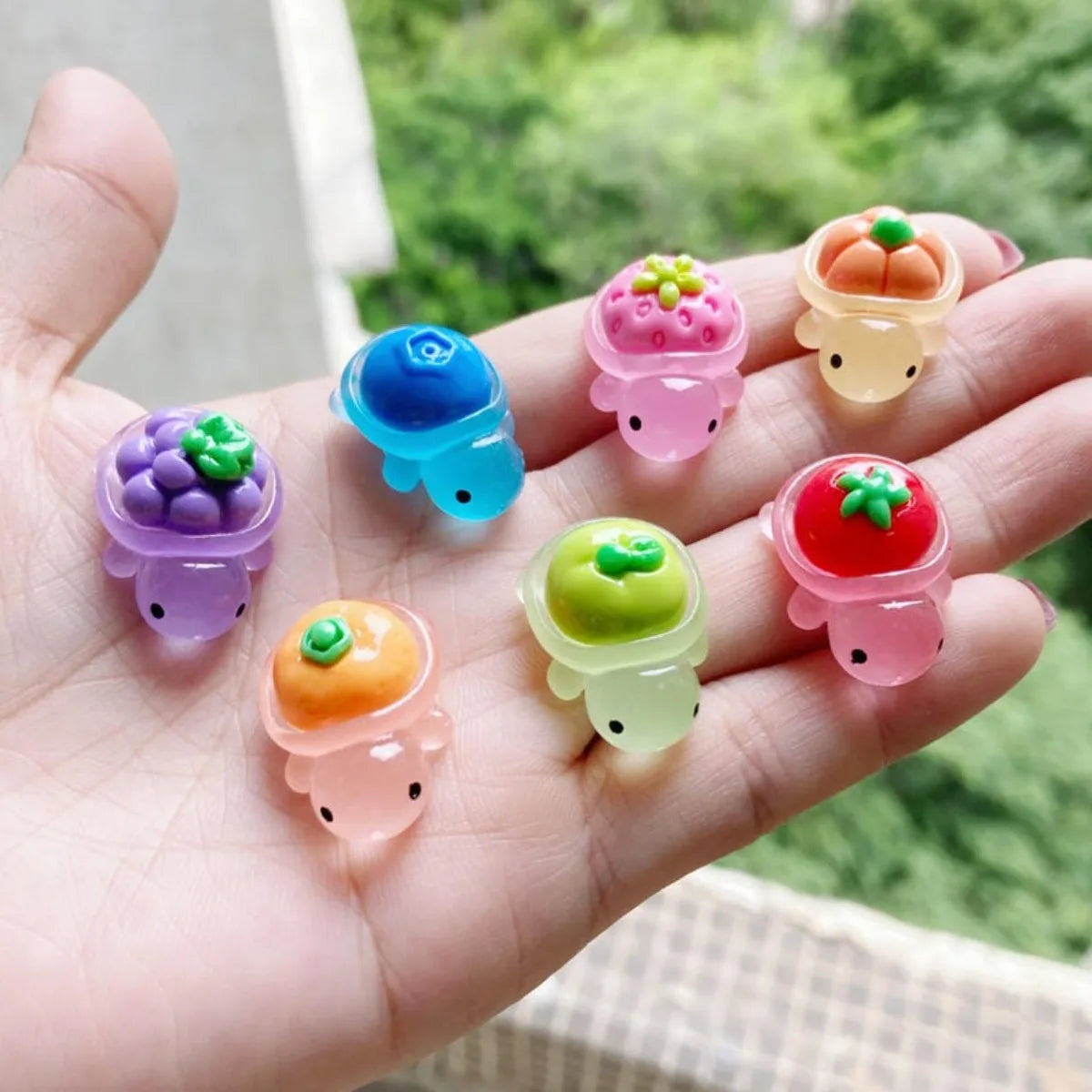 A hand holds several glow-in-the-dark fruit turtle resin charms in various colors like pink, purple, green, and orange.