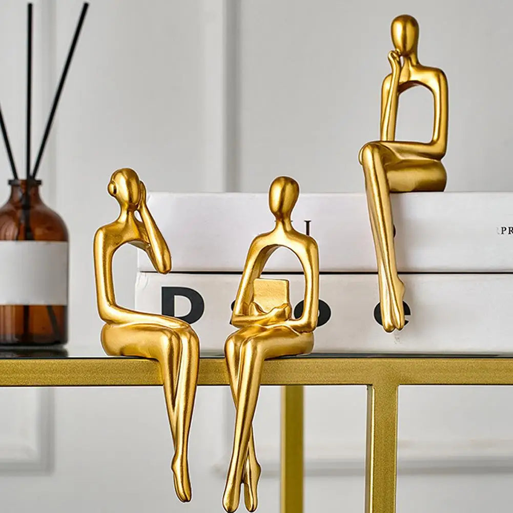 Three gold abstract thinker sculptures sitting on a glass desk with stacked books.