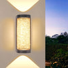 A vertical, cylindrical LED crystal outdoor wall sconce with a warm glow mounted on a light-colored wall.