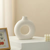 A white ceramic vase shaped like a donut sits on a light surface next to a framed sign.