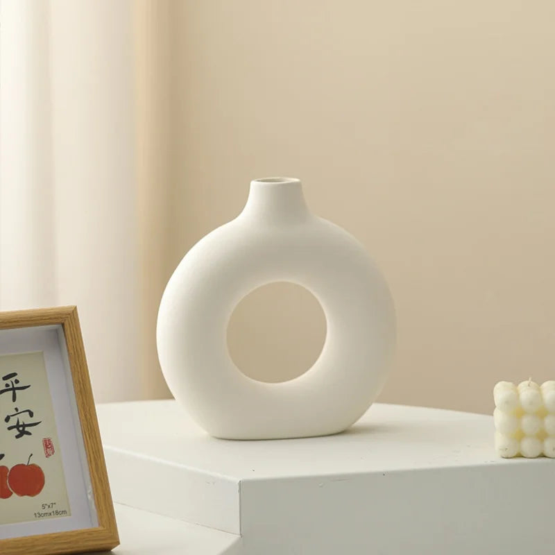 A white ceramic vase shaped like a donut sits on a light surface next to a framed sign.