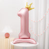 A pink foil balloon shaped like the number one with a gold crown on top, displayed on a white table.