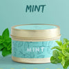 A tin of mint-scented soy wax travel candle with fresh mint leaves surrounding it.