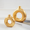 A golden, smooth, geometric resin vase with a rounded neck and ring shape on a white surface.