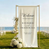 A white linen banner with gold frame and elegant script welcoming guests to a wedding, set outdoors near the sea.