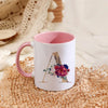 A white ceramic coffee mug with a pink handle featuring a gold letter initial decorated with colorful flowers.