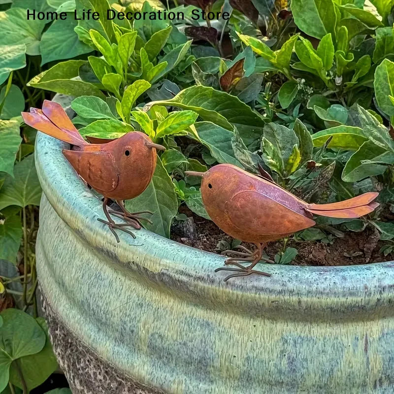 Two rustic metal bird sculptures in shades of brown and orange perched on a green plant in a terracotta pot.