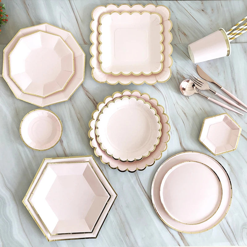 A collection of gold and pink disposable party tableware including plates, cups, napkins, and straws arranged on a light surf