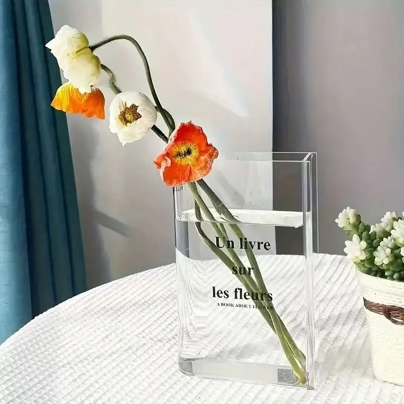 A clear acrylic book vase holds a single orange and white flower arrangement on a white cloth.