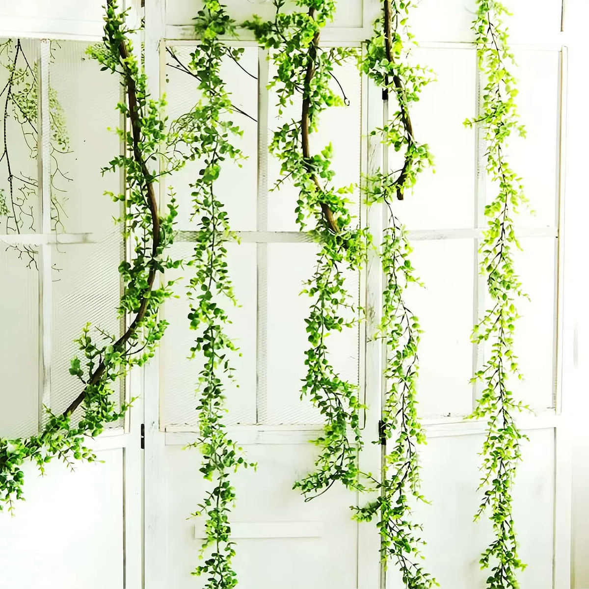 A vertical display of artificial simulation vine garlands with bright green leaves hanging against a white wooden background.