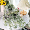 Plantes d'eucalyptus artificielles suspendues sur une table blanche à côté d'une lampe de couleur crème.