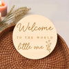 A round wooden sign with gold lettering that reads Welcome to the world little one resting on a woven mat next to a candle an