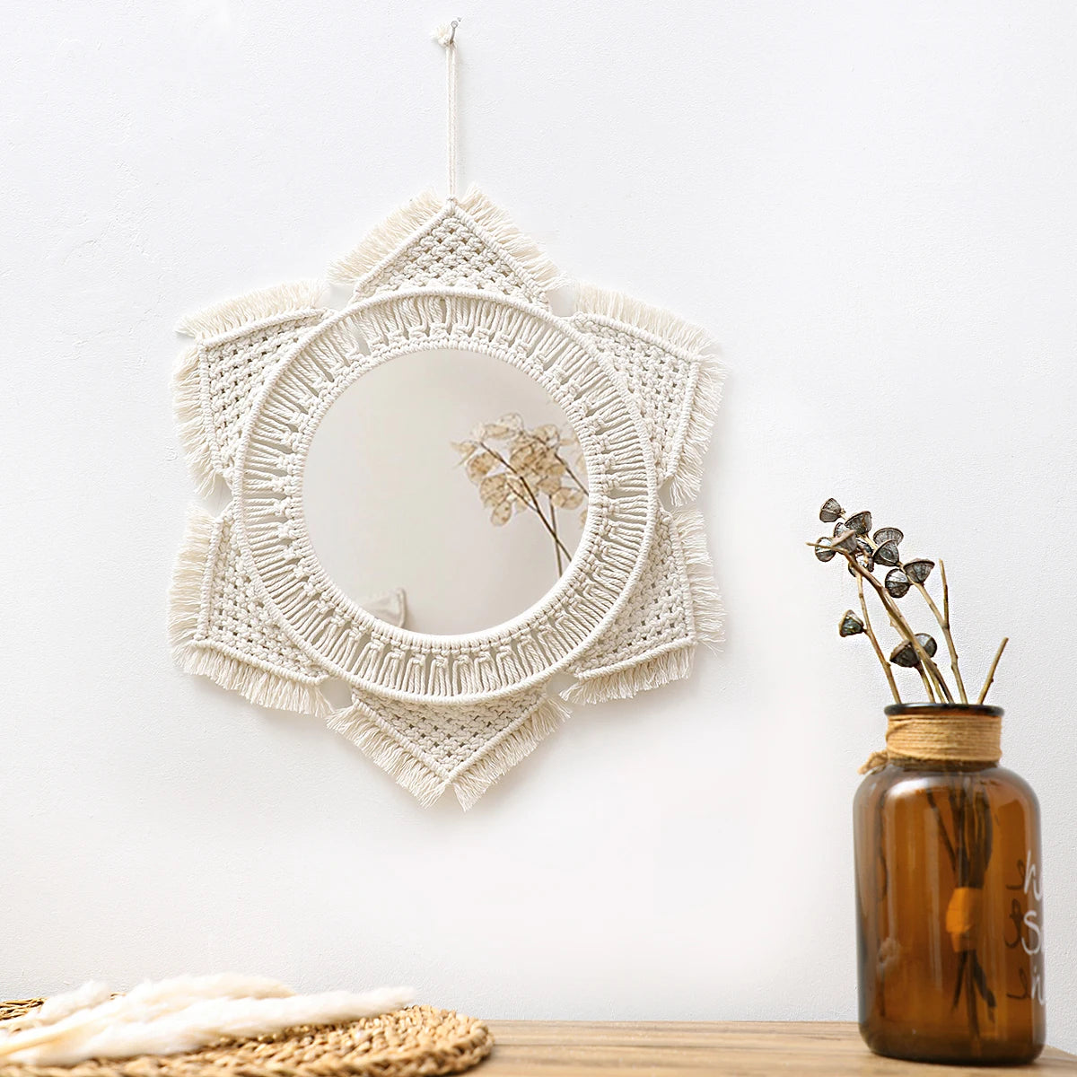 A white macrame round wall mirror with fringe, hanging on a white wall next to a glass vase with dried flowers.