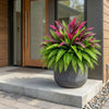 A potted artificial Dracaena Boston fern with vibrant magenta and green leaves sits on a concrete step by a modern glass door