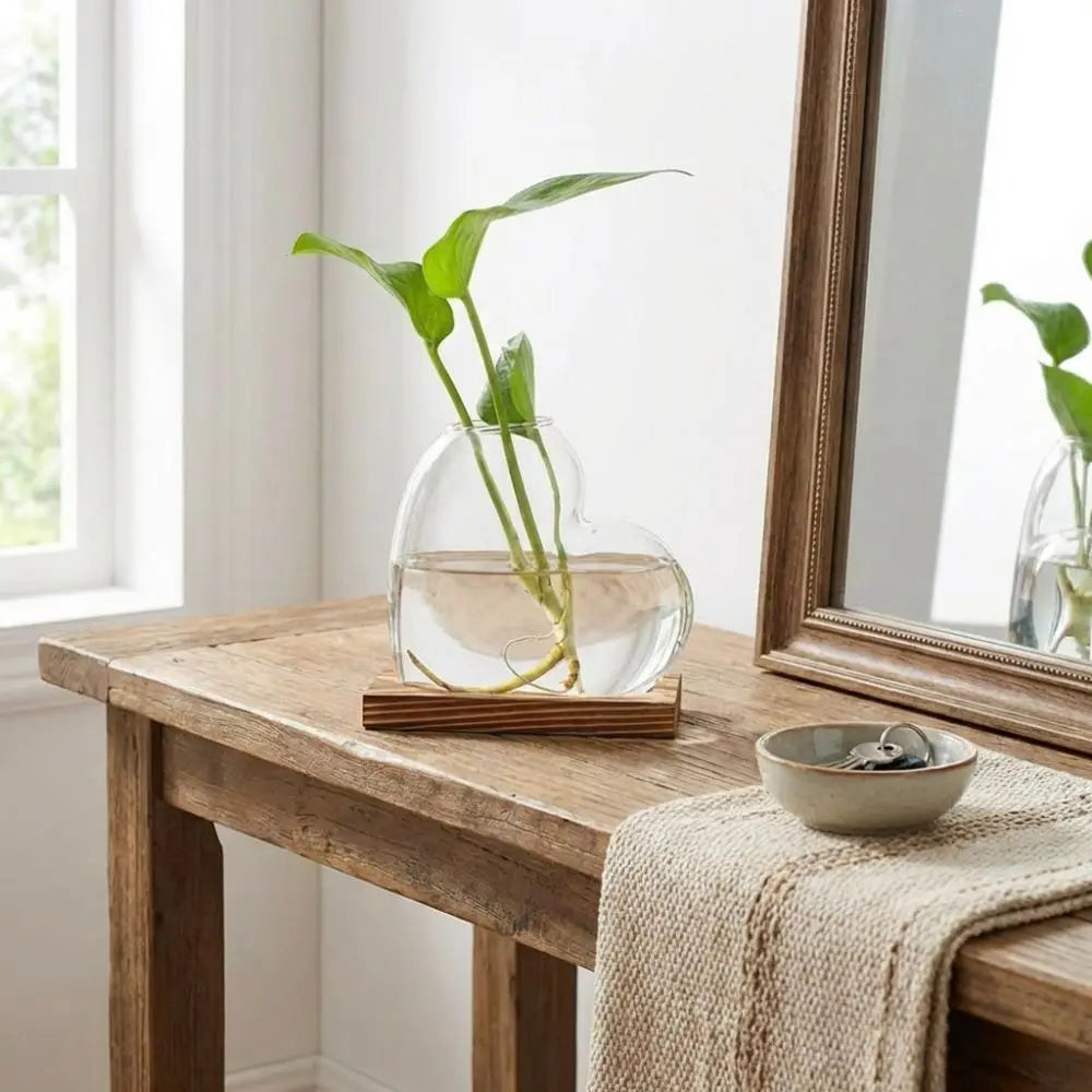 A clear glass vase holding a green plant in a heart shape rests on a wooden stand on a rustic table.