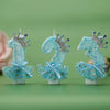 Three glittery blue birthday candles with crown toppers and sheer fabric skirts