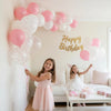 Two young girls smiling while holding a large arrangement of pink and white balloons in a bright room.