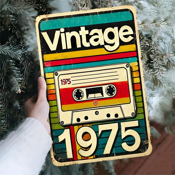 A vintage style metal wall sign featuring the year 1975 on a colorful background, held by a hand against a green pine branch.