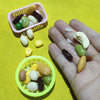 A hand holds several realistic resin fake corn miniatures in various colors and shapes, arranged in small plastic baskets.