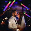 A couple is embraced while being illuminated by colorful LED foam glow sticks in a dark setting.