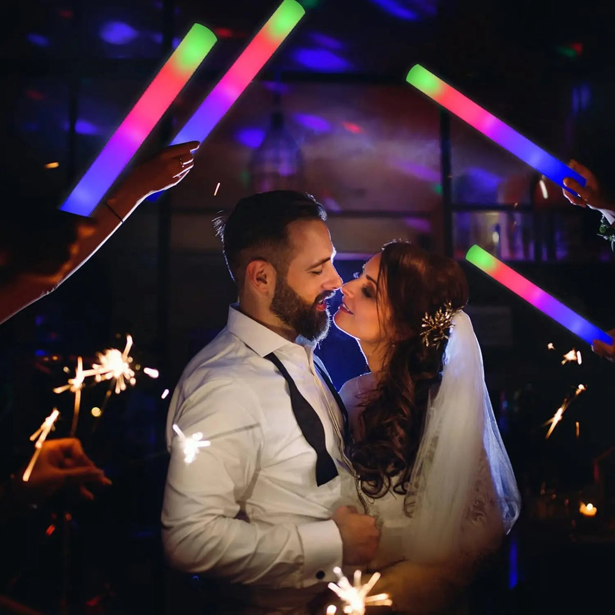 A couple is embraced while being illuminated by colorful LED foam glow sticks in a dark setting.