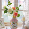A pink and white ceramic bird floral vase sits on a table next to an open book and sheer fabric.