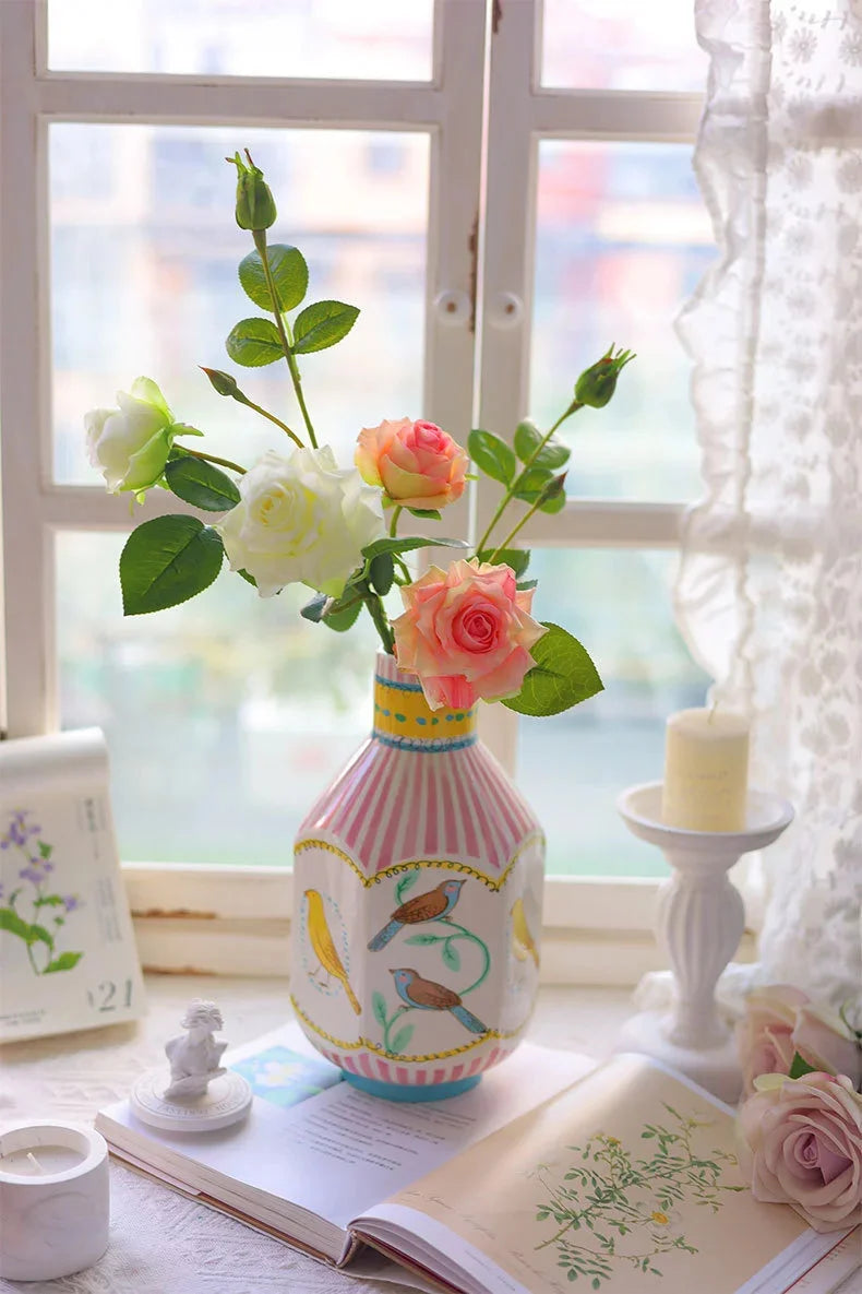 A pink and white ceramic bird floral vase sits on a table next to an open book and sheer fabric.