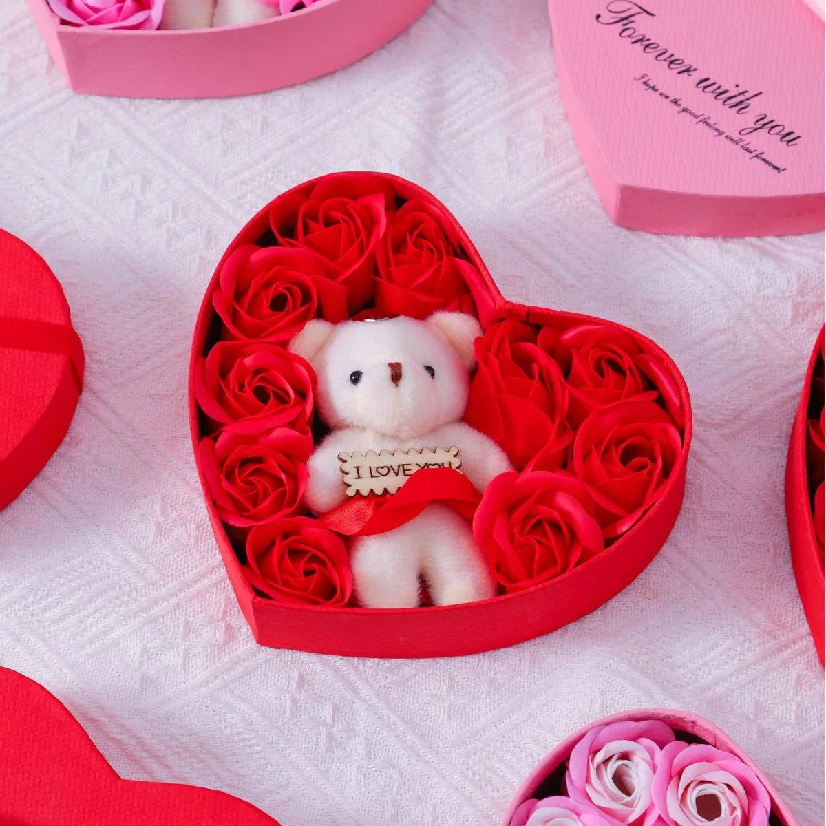 A red gift box filled with red roses and a white bear soap, set on a pink quilted fabric.