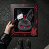 A dark gray bunny plush keychain is displayed inside its packaging with red lightning bolt accents.