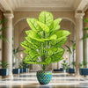 A large artificial floor plant with vibrant green leaves sits in a patterned ceramic pot on a polished wooden floor.