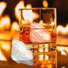 A glass of amber liquid with a large pink ice crystal and a clear ice shard resting on a reflective surface.