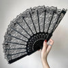 A hand holding an elegant black lace folding fan with a scalloped edge and decorative accents.