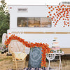 A white RV is decorated with an orange artificial maple leaf garland hanging vine and a rustic wooden sign.