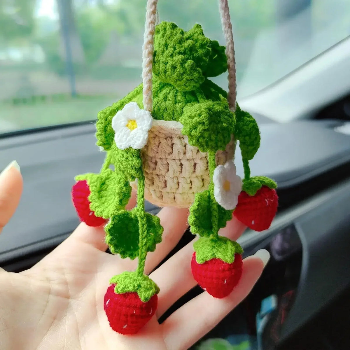 Hand-knitted hanging plant basket decor featuring green leaves and white flowers, displayed in a hand.