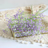A bouquet of purple artificial gypsophila flowers in a gold frame rests on a beige linen fabric.