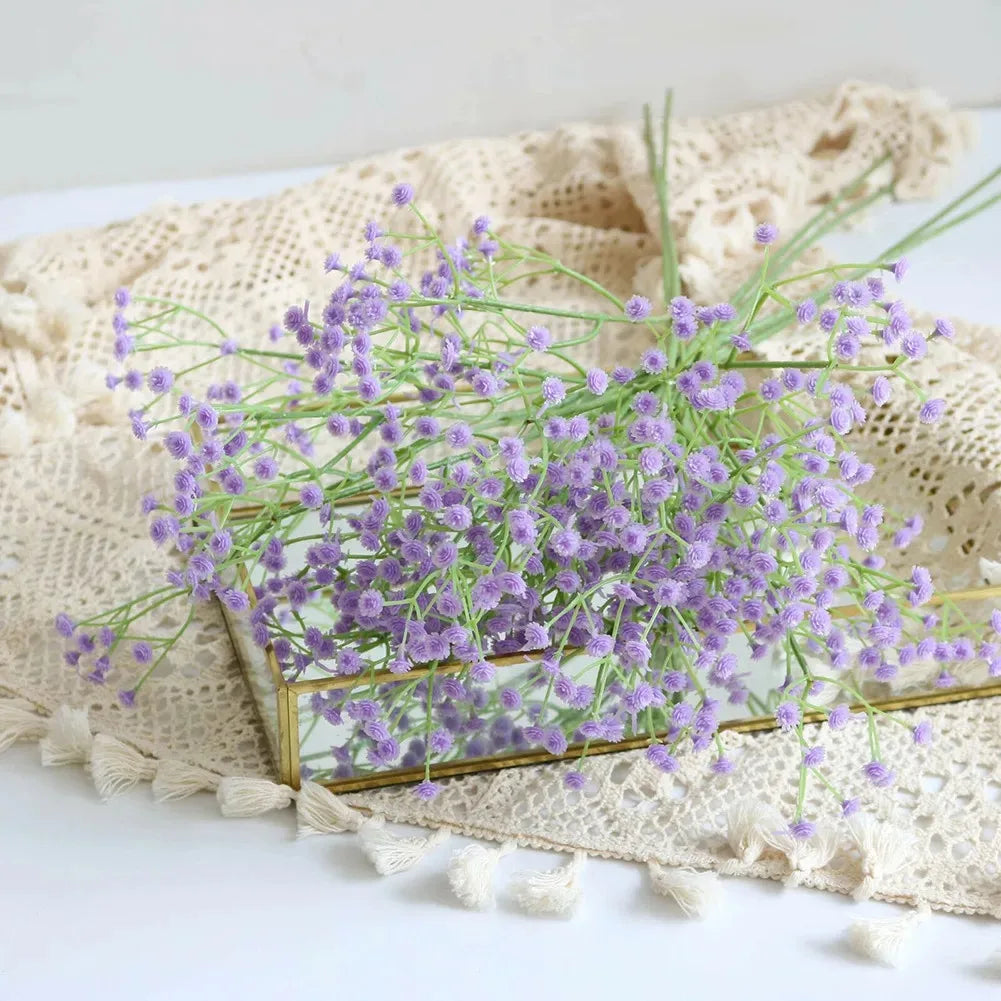 A bouquet of purple artificial gypsophila flowers in a gold frame rests on a beige linen fabric.