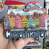 A hand holds a collection of wooden fridge magnets depicting colorful Scandinavian-style houses and clouds, with the word Cop