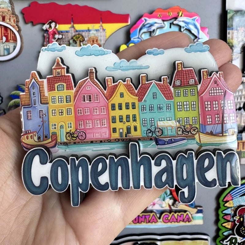 A hand holds a collection of wooden fridge magnets depicting colorful Scandinavian-style houses and clouds, with the word Cop