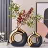 A pair of black and gold ceramic flower vases with green foliage are displayed on a white surface.