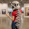 A woman wearing a white dinosaur skeleton plush costume, featuring a skull head and a full spine, stands in an art gallery.