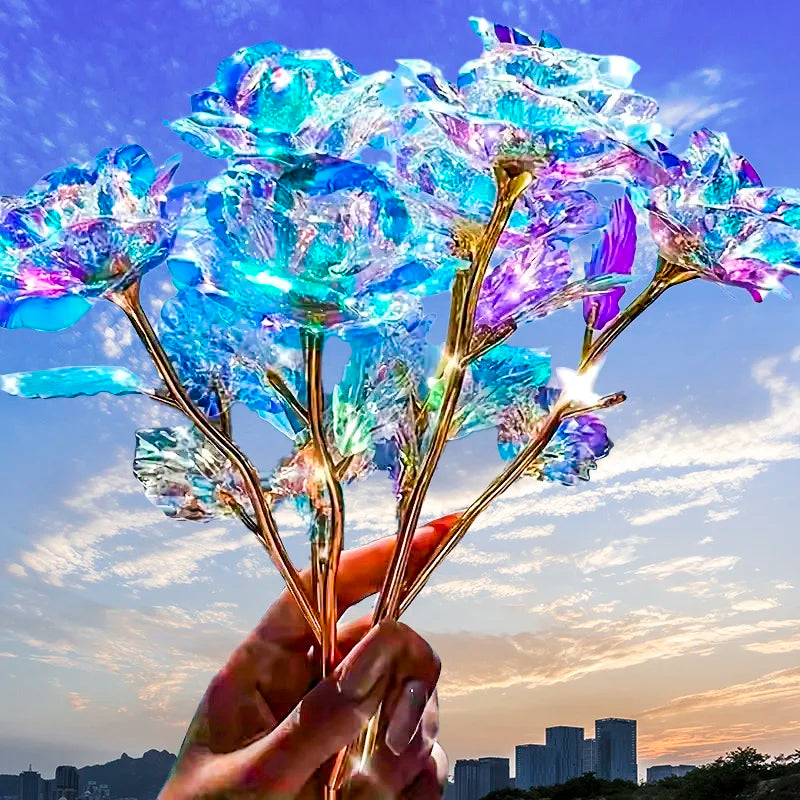 A hand holds a vibrant iridescent gold foil rose with blue and purple petals against a sunset sky.