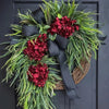 A rustic wreath of green fir and deep red hydrangeas tied with a black ribbon, displayed on a dark door.